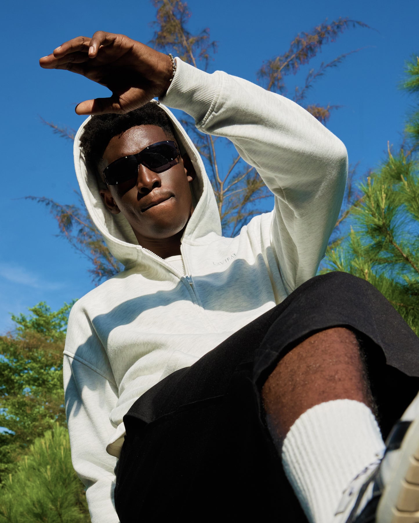 Back view of a man wearing the white LAVIEM Boxy Double-Zip Hoodie with sunglasses, styled with cotton cargo short and sneakers in an urban streetwear setting.